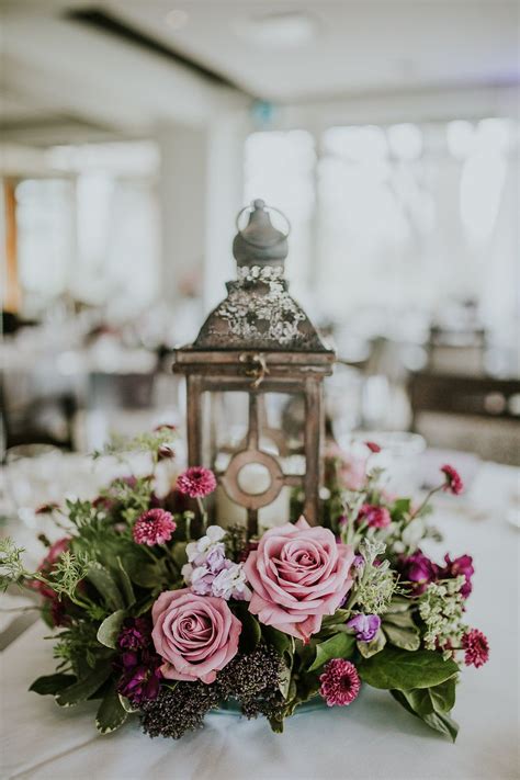 A Lantern With Flowers And Greenery Sits On A Table At A Wedding In A