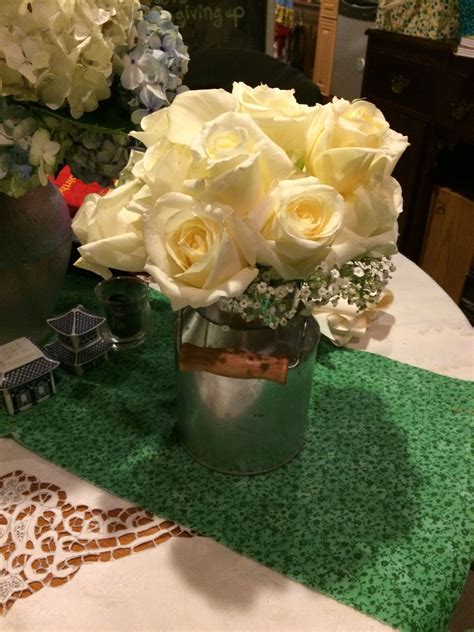 White Roses Are In A Silver Vase On A Green Table Cloth Near Other