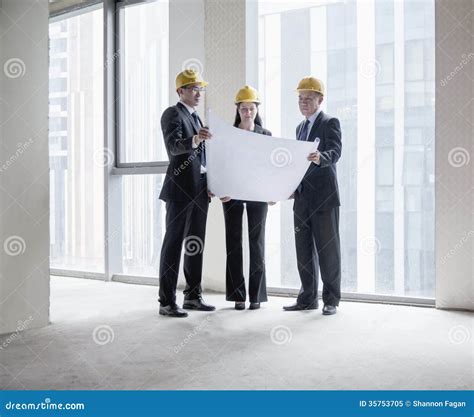 Three Architects In Hardhats Examining A Blueprint In An Office