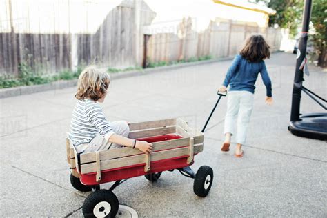 Rear View Of Boy Pulling Brother In Toy Wagon At Yard Stock Photo