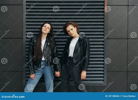Street Portrait Of Two Stylish Girls In Leather Jackets Are Standing Against A Dark Wall