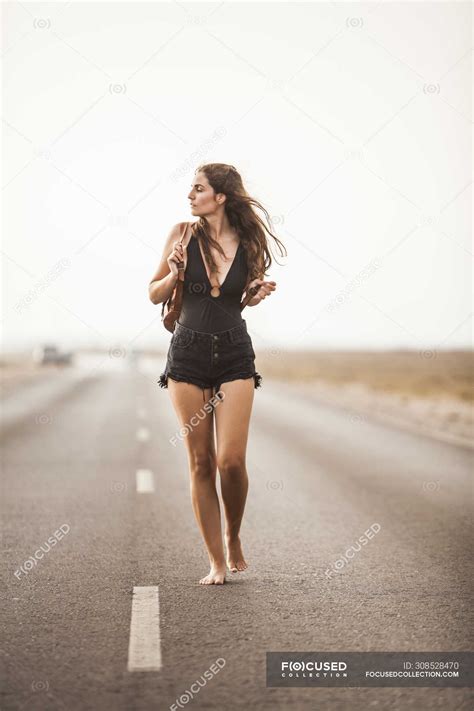 Attractive Young Barefoot Woman Walking On Empty Road Holding Backpack