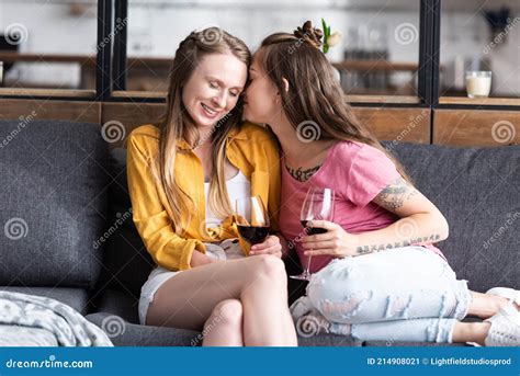 Two Lesbians Holding Wine Glasses And Kissing While Sitting On Sofa In Living Room Stock Image