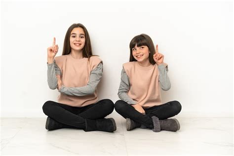 irmãs mais novas sentadas no chão isoladas no fundo branco mostrando e levantando um dedo em