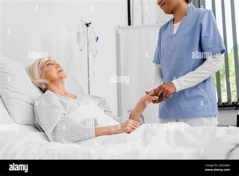 African American Nurse Holding Hand Of Senior Patient With Nasal Cannula On Hospital Bed Stock