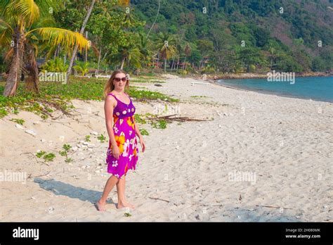 A Mature Woman With Long Blond Hair Walks Along A Sandy Beach Among Palm Trees On A Tropical