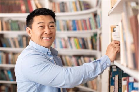 Premium Photo Cheerful Asian Man Taking Book From Bookshelf In Modern
