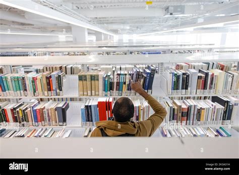 Male College Student Reaching For Book On Shelf In Library Stock Photo