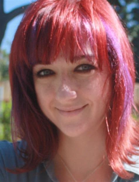 A Young Woman With Red Hair And Blue Eyes Smiles At The Camera While Wearing A Gray Shirt