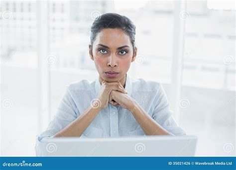 Stern Businesswoman Sitting At Her Desk Looking At Camera Stock Photo