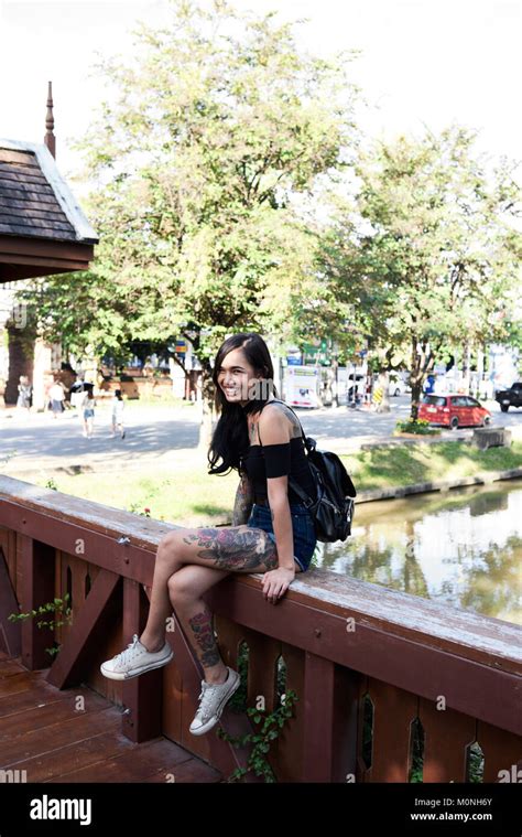 Tattooed Asian Brunnete Girl Smiling At Camera While Sitted On A Wooden Bridge In Summer Stock