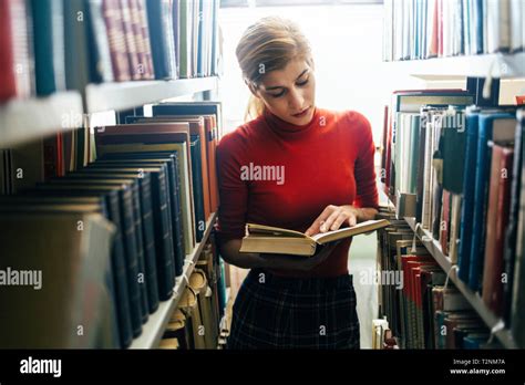 Young Attractive Librarian Reading A Book Between Library Bookshelves