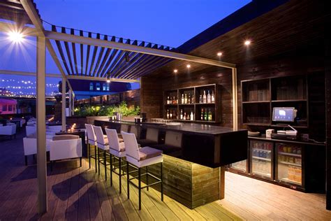 An Outdoor Bar With White Chairs And Wooden Flooring At Night Lit Up