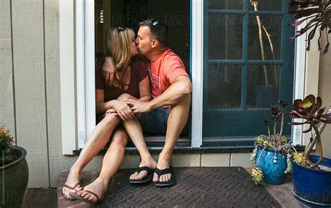 Married Middle Aged Couple Sitting In The Doorway Kissing By Stocksy