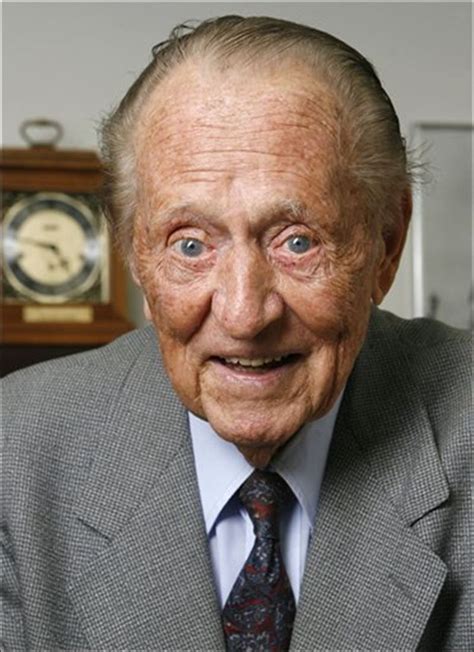 An Old Man In A Suit And Tie Smiling At The Camera With A Clock Behind Him