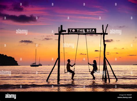 Romantic Couple In Love Sitting Together On Rope Swing At Sunset Beach Silhouettes Of Young Man