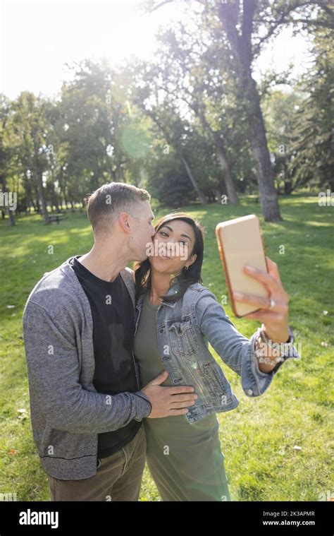 Happy Affectionate Couple Kissing And Taking Selfie In Sunny Park Stock