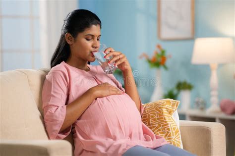 indian pregnant woman drinking water while sitting on sofa at home concept of hydration