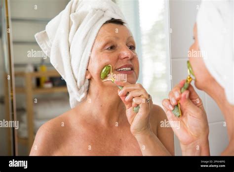 Mature Beautiful Woman Doing Facial Massage With Jade Roller Looking In The Mirror In Bathroom