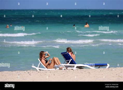 Two Girls In Bikini Tanning On Deck Chairs On Sea Waves Background Beach With White Sand On
