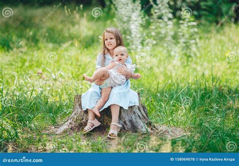 Two Little Sisters Poses Outdoors While Sitting On Wooden Stump In