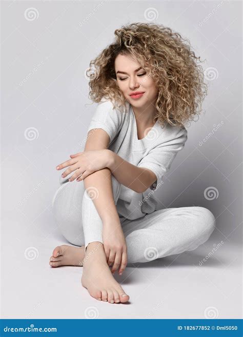 Blonde Curly Woman In White Suit Sitting On The Floor With Crossed Hands Looking Down Shyly