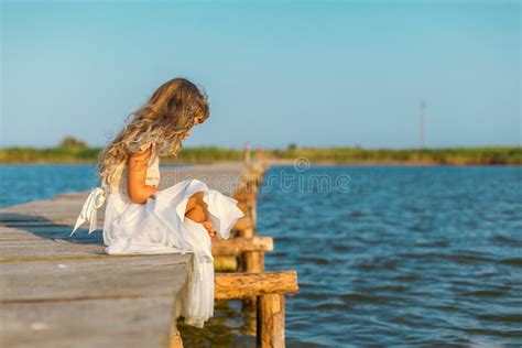 Little Girl With Long Blond Hair Sitting On The Pier Stock Photo