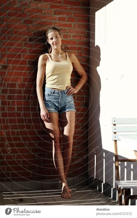Young Tall Blonde Woman Standing On Balcony Barefoot In The Sun Leaning Against Brick Wall And