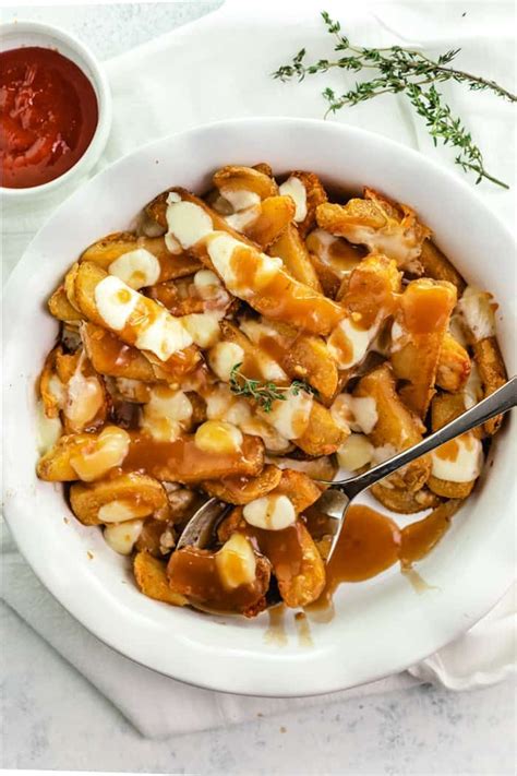 Overhead View Of A White Bowl Of Poutine Next To Ketchup And Rosemary
