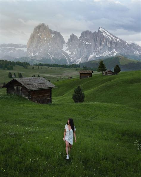 A Woman In A White Dress Is Walking Through The Grass With Mountains In