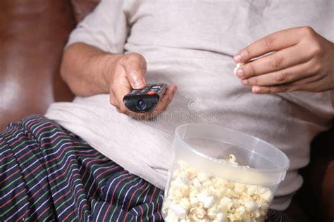 A Fat Man Eating Popcorn And Holding Tv Remote Sitting On Sofa Stock