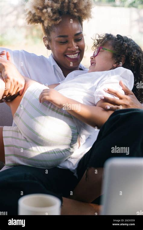 African American Woman Tickling Adorable Daughter While Lying On Couch In Courtyard And Having