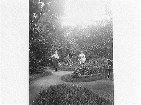 två stående kvinnor och en sittande man i en prydlig trädgård med anlagda grusgångar rabatter
