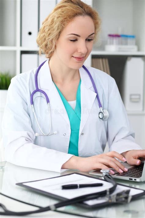 happy blonde female doctor sitting at the table and typing by laptop computer medicine