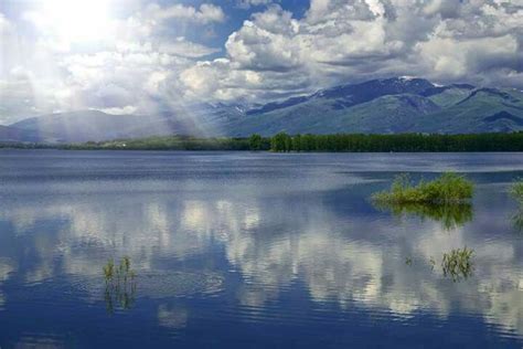 The Sun Shines Brightly Through Clouds Over A Lake With Mountains In