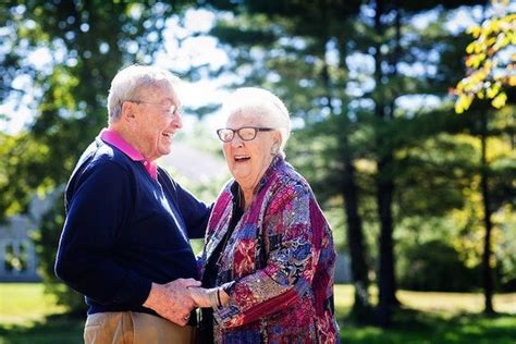 An Older Man And Woman Standing Next To Each Other In The Grass With Trees Behind Them