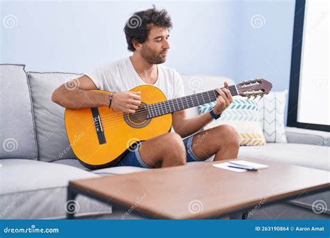 Young Hispanic Man Playing Classical Guitar Sitting On Sofa At Home