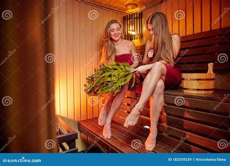 Two Beautiful Girls With Broom On Wooden Bench At Sauna In Steam Room