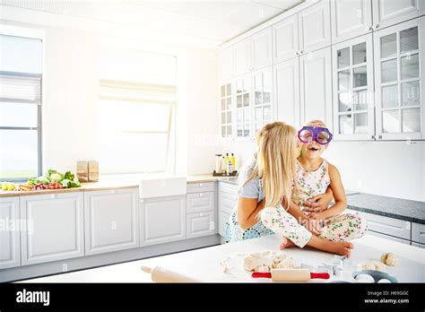 Blond Haired Mother Kissing Daughter On Cheek In Kitchen Beside Bread