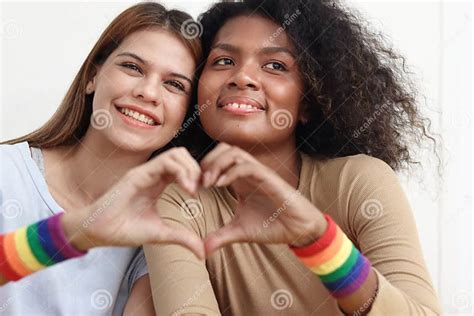 happy smiling homosexual lesbian couple with rainbow flag wristband making heart sign by hand