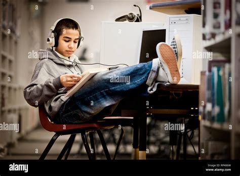 Teenage Boy Sitting With His Feet Up And Reading A Book With Headphones