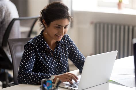 Smiling Indian Female Employee Using Laptop At Workplace Helpside