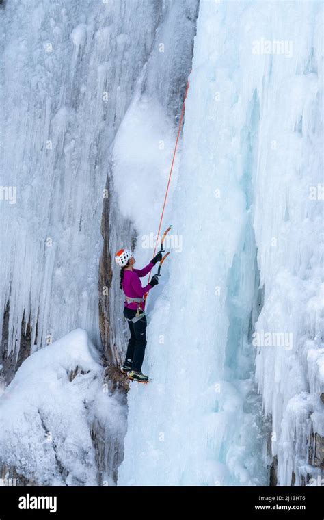 A Female Ice Climber Ascending An Ice Wall Using Ice Axes And Crampons