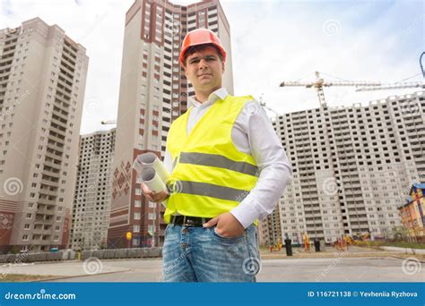 Portrait Of Smiling Male Engineer In Hardhat Standing With Blueprints