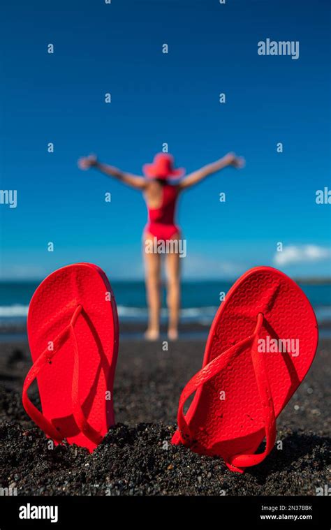 Red Flip Flops In The Black Sand And Silhouette Of Woman In Red Swimsuit Standing On The Ocean