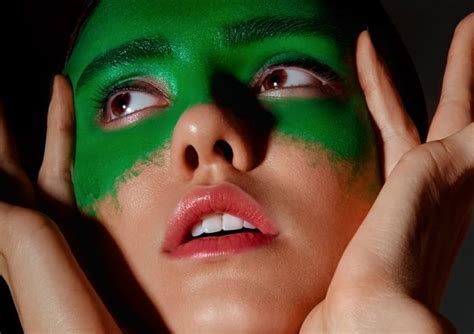 A Woman With Green Paint On Her Face And Hands Around Her Head Posing