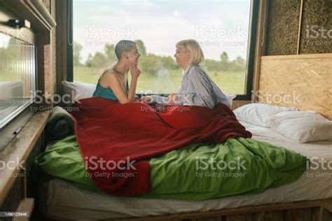 Two Lesbian Women Dressed In Pajamas Sit In Bed By The Window In A Glamping Cabin And Enjoy