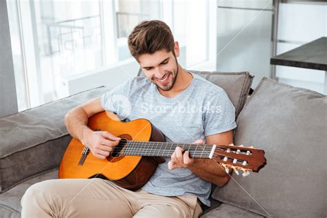 Happy Man Playing On The Guitar While Sitting On Sofa At Home Looking