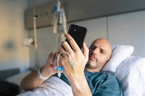 Male Patient Using Mobile Phone While Lying On Bed In Hospital Ward