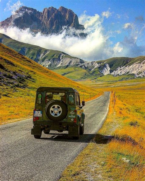 A Jeep Driving Down The Road In Front Of A Mountain Range With Clouds
